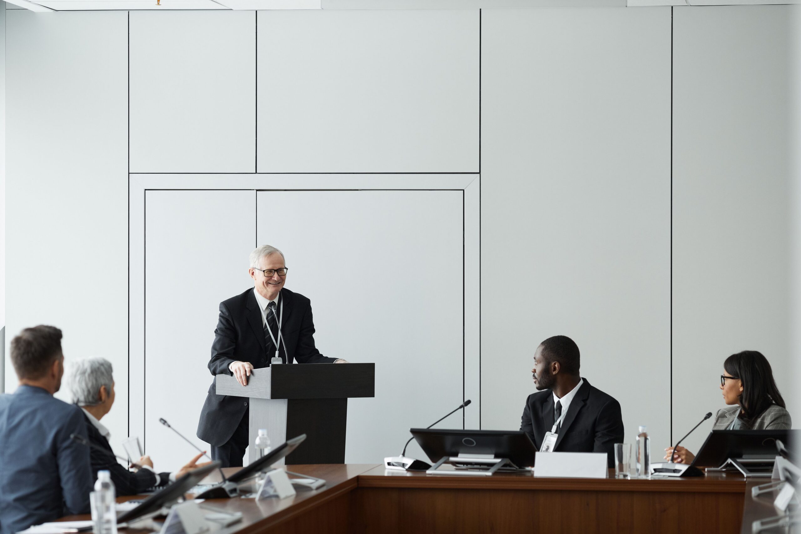 Formal business meeting with a diverse group and speaker at a podium in a conference room.